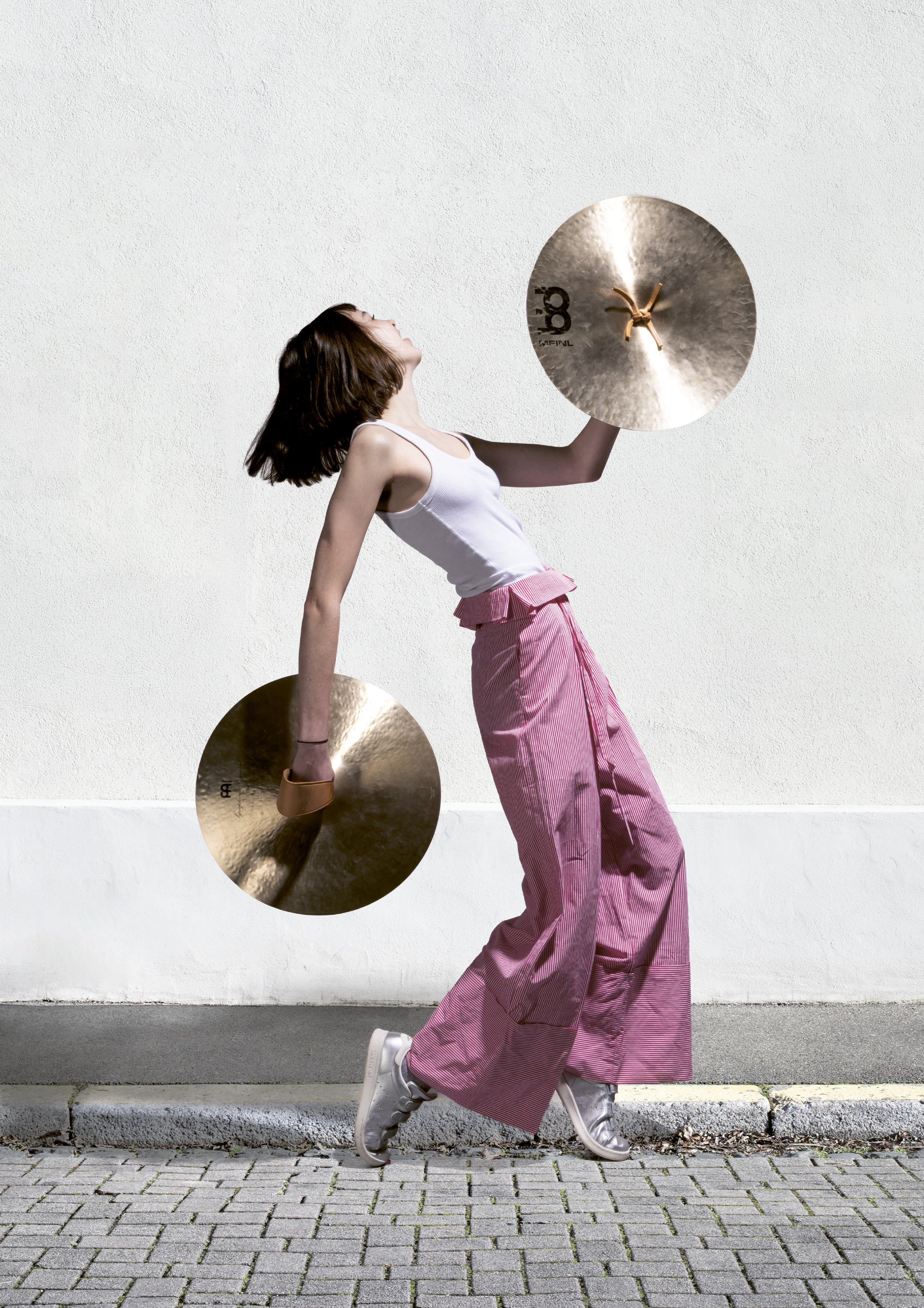 Femme en mouvement tenant deux cymbales, l’une devant elle et l’autre derrière, sur fond de mur blanc, dans une posture dynamique évoquant la musique et le rythme